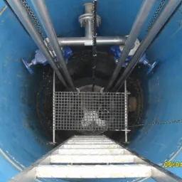 Wnętrze studni z niebieskimi ścianami, widoczna drabina schodząca w d&oacute;ł, rury z zaworami, metalowa krata i łańcuchy.