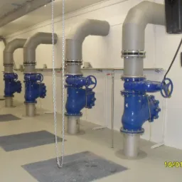 Rząd niebieskich zawor&oacute;w przemysłowych z szarymi rurami w sterylnej hali, widoczne kraty podłogowe i łańcuch zwisający z sufitu, oświetlenie jarzeniowe.