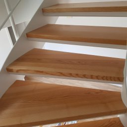 Darma - Jasne, drewniane schody z białymi metalowymi elementami konstrukcyjnymi, widziane z dołu, ukazujące naturalny rysunek drewna i nowoczesny design.