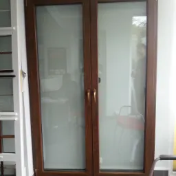 Dwuskrzydłowe drzwi balkonowe z brązową ramą i złotymi klamkami, widok na wnętrze z osobą przy biurku i drugą osobą stojącą.