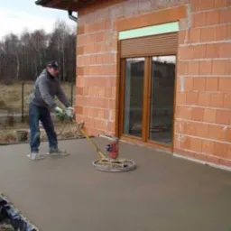 Pracownik w kombinezonie i ochronnych butach wygładza świeżo wylaną posadzkę maszyną z obrotowymi dyskami na tle surowego budynku z cegły.