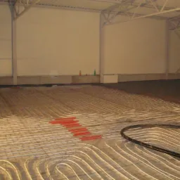 Instalacja ogrzewania podłogowego z rur w dużej hali, widoczne czerwone elementy mocujące i ciemna wylewka na obrzeżach.