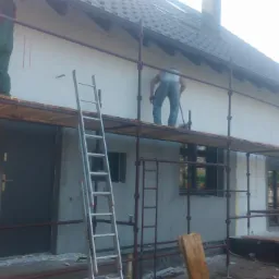 Prace na rusztowaniu przy ocieplaniu ściany domu jednorodzinnego, widoczne płyty styropianowe na elewacji, drabina oparta o ścianę, w tle dach z ciemnej dachówki.