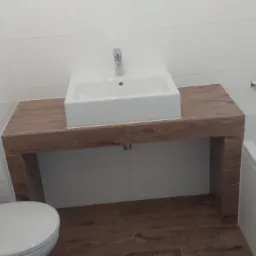 Biała, prostokątna umywalka nablatowa z chromowaną baterią na blacie z drewnopodobnych płytek, obok wanna i toaleta, białe płytki na ścianach.