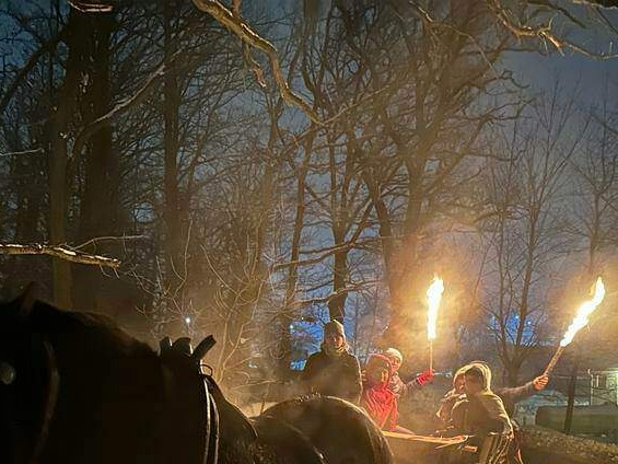 Nocny przejazd bryczką przez ośnieżony las, grupa osób trzymająca płonące pochodnie, widok na zaprzęg konny w ciemności.