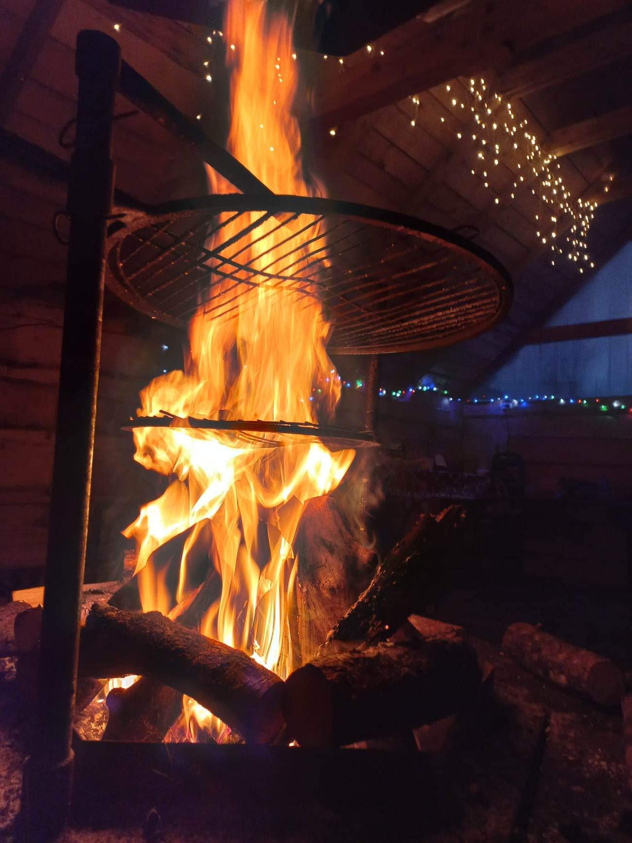 Ognisko pod rusztem w zadaszonym miejscu, udekorowane lampkami, płomienie oświetlają drewnianą konstrukcję w Miłkowie.