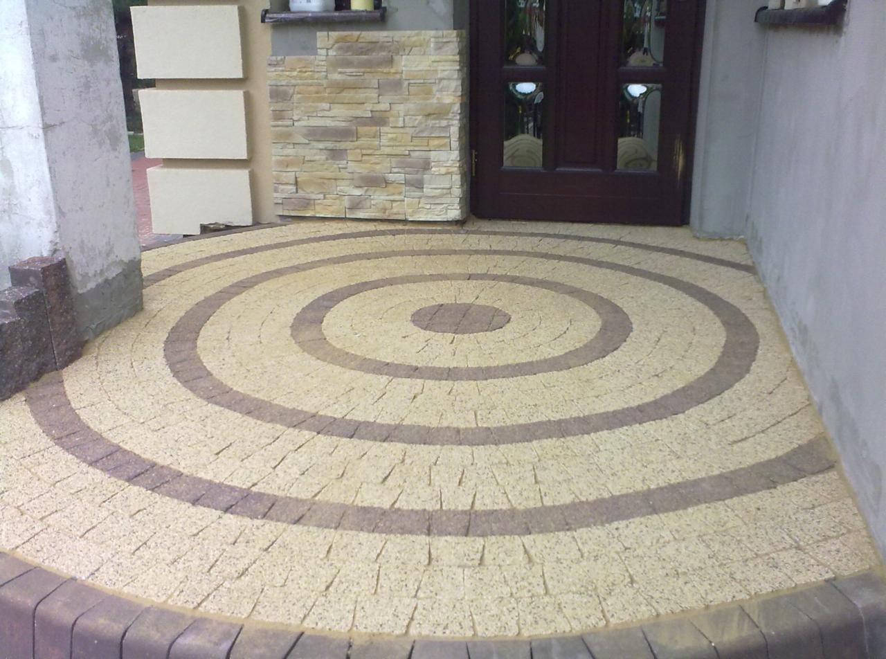 Okrągły wzór z kostki brukowej w odcieniach beżu i brązu, ułożony przed wejściem do budynku. Widoczne elementy elewacji z kamienia dekoracyjnego.