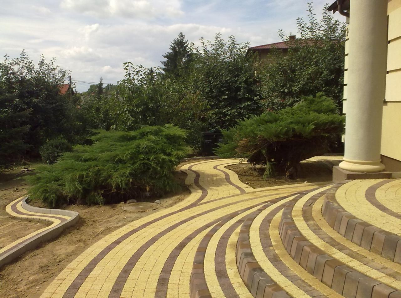 Wzorzysta ścieżka z kostki brukowej w ogrodzie, prowadząca od schodów z kolumną w tle, z elementami zieleni. Widok na posesję z roślinnością.