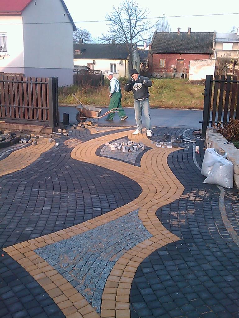 Ułożona kostka brukowa w oryginalny wzór na podjeździe, z pracownikami w tle. Widoczne różne kolory kostki i elementy dekoracyjne.