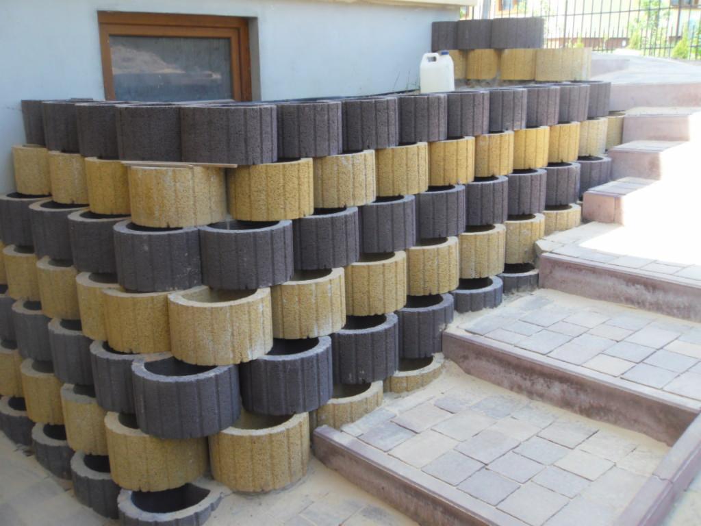 Konstrukcja z betonowych donic w odcieniach szarości i beżu, tworząca mur oporowy przy schodach z kostki brukowej, z widocznym oknem w tle.