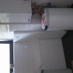 Biały bojler i piec gazowy wiszą na ścianie w łazience z ciemną podłogą. Widoczne rury i armatura. Drzwi w tle. Ujęcie z dołu.