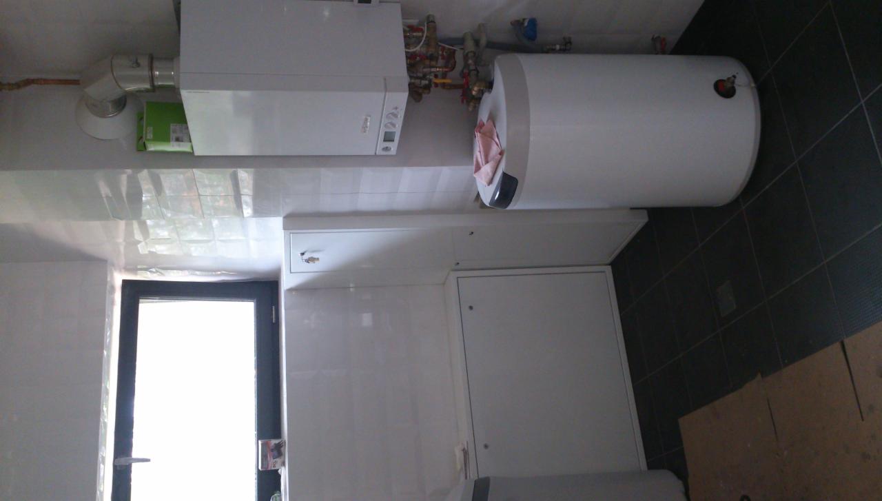 Biały bojler i piec gazowy wiszą na ścianie w łazience z ciemną podłogą. Widoczne rury i armatura. Drzwi w tle. Ujęcie z dołu.