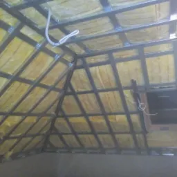 Żółta wełna mineralna w konstrukcji metalowej na poddaszu, widoczny kabel elektryczny i fragment okna dachowego.