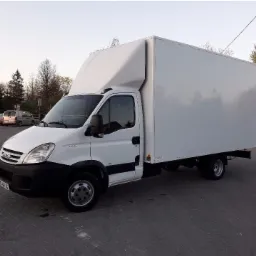 Biały samochód dostawczy Iveco z zabudową kontenerową, zaparkowany na kostce brukowej, widoczne drzewa w tle.