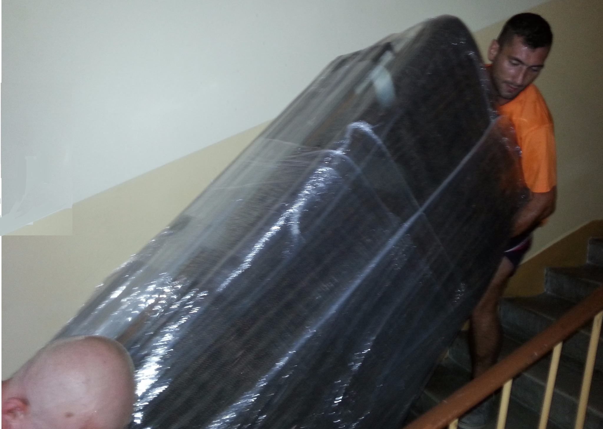 Mężczyzna w pomarańczowej koszulce wnosi po schodach duży, owinięty folią mebel.