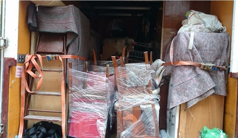 Wnętrze załadowanej ciężarówki: meble owinięte folią i kocami zabezpieczone pasami transportowymi, widoczna drabina i torby z rzeczami.