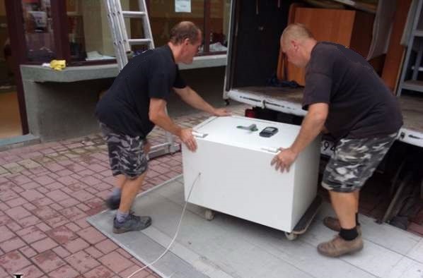 Dwóch mężczyzn w czarnych koszulkach i moro spodenkach przenosi białą skrzynię na kółkach, prawdopodobnie lodówkę, z chodnika do wnętrza furgonetki, na skrzyni leżą klucze i telefon.