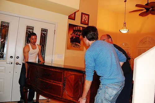 Trzech mężczyzn w trakcie wnoszenia ciężkiej, drewnianej komody z czarnym blatem do jasnego wnętrza domu. Jeden z mężczyzn ma na sobie biały podkoszulek.