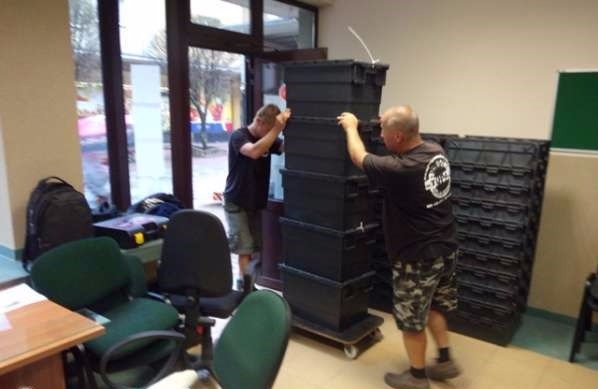 Dwóch mężczyzn w Elblągu układa wieżę z czarnych plastikowych skrzyń na wózku transportowym, obok sterta identycznych skrzyń.