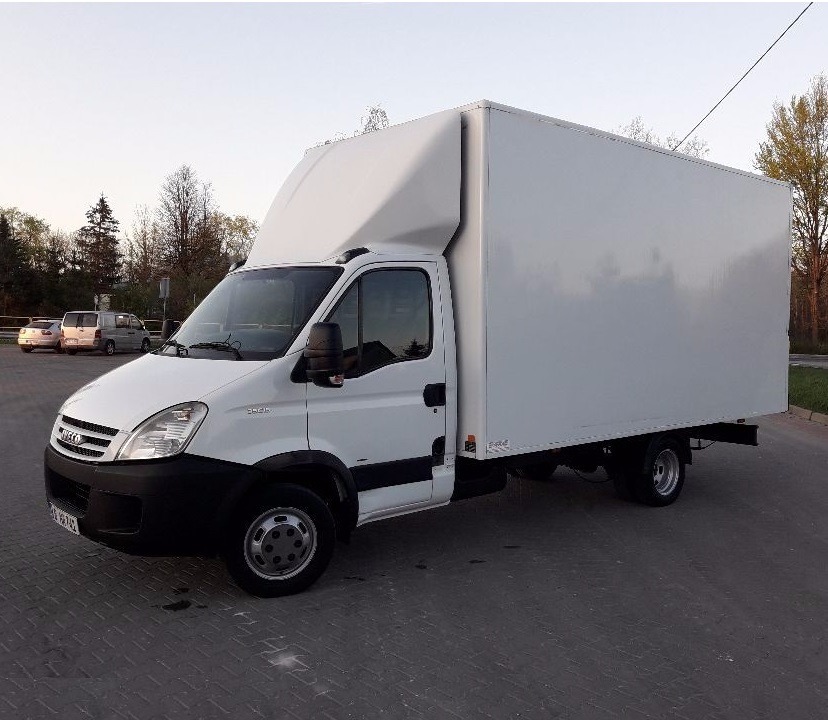 Biały samochód dostawczy Iveco z zabudową kontenerową, zaparkowany na kostce brukowej, widoczne drzewa w tle.