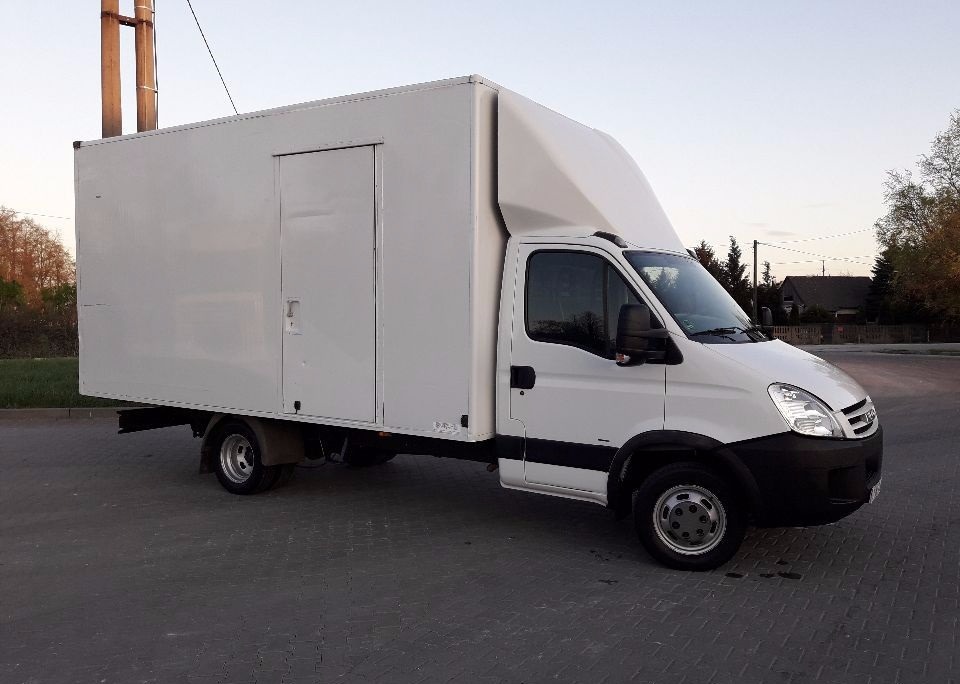 Biały samochód dostawczy z zabudową kontenerową, zaparkowany na kostce brukowej, widok z boku, z drzwiami z tyłu kontenera.