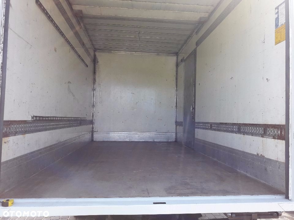 Wnętrze paki busa transportowego z metalowymi szynami mocującymi na ścianach, widok od tyłu, pusta przestrzeń ładunkowa z lekko zabrudzoną podłogą.