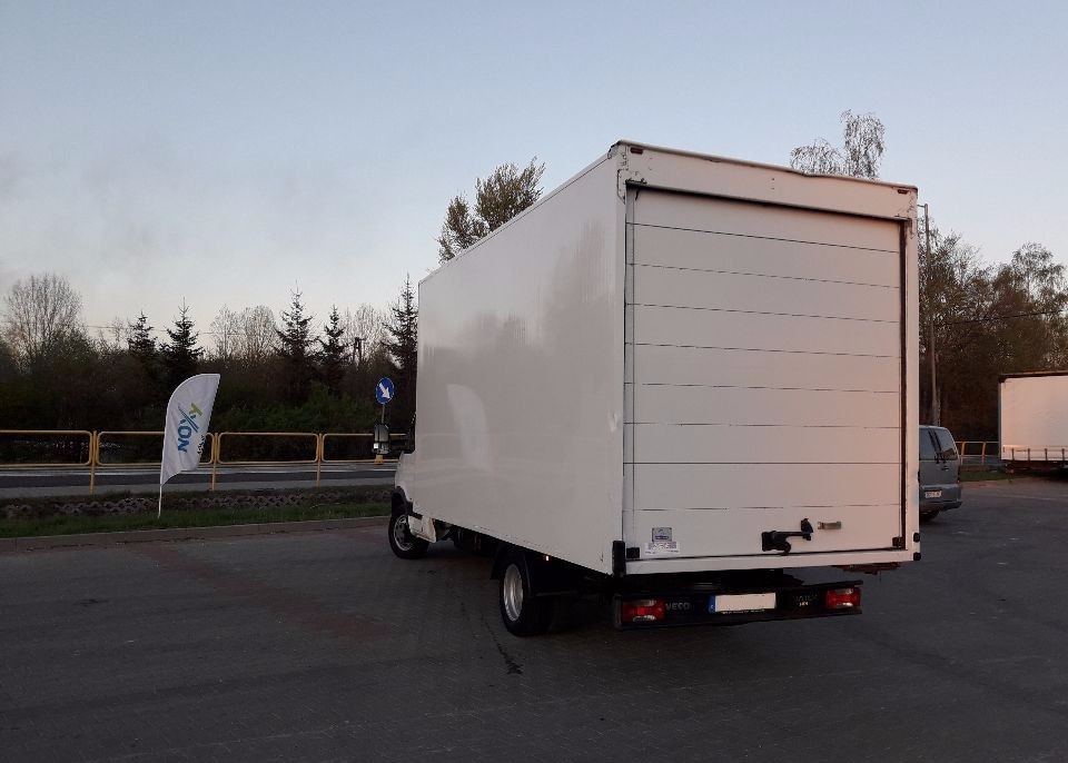 Biały samochód dostawczy z zamkniętymi drzwiami rolowanymi, widziany z tyłu na parkingu, obok drogi z flagą reklamową i znakiem drogowym.