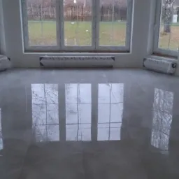 Błyszcząca, szara posadzka w nowym pomieszczeniu z dużymi oknami, grzejniki zabezpieczone folią, widoczne odbicie drzew w oknach.