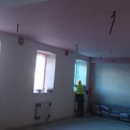 Pracownik w ż&oacute;łtej kamizelce i kasku na drabinie, montaż płyty kartonowo-gipsowej na suficie w pomieszczeniu z trzema oknami, widoczne kable zwisające z sufitu i worki z materiałami budowlanymi...