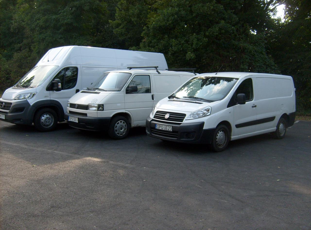 Trzy białe busy dostawcze różnych marek zaparkowane obok siebie na asfaltowym placu, w tle drzewa. Widoczne tablice rejestracyjne.