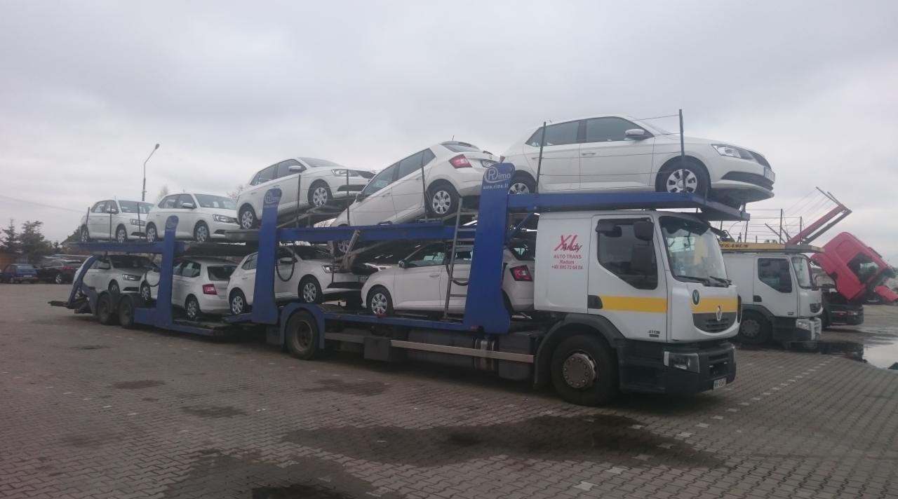 Laweta przewożąca osiem białych samochodów marki Skoda Fabia na parkingu, widoczne logo firmy transportowej Xlay Auto Trans.