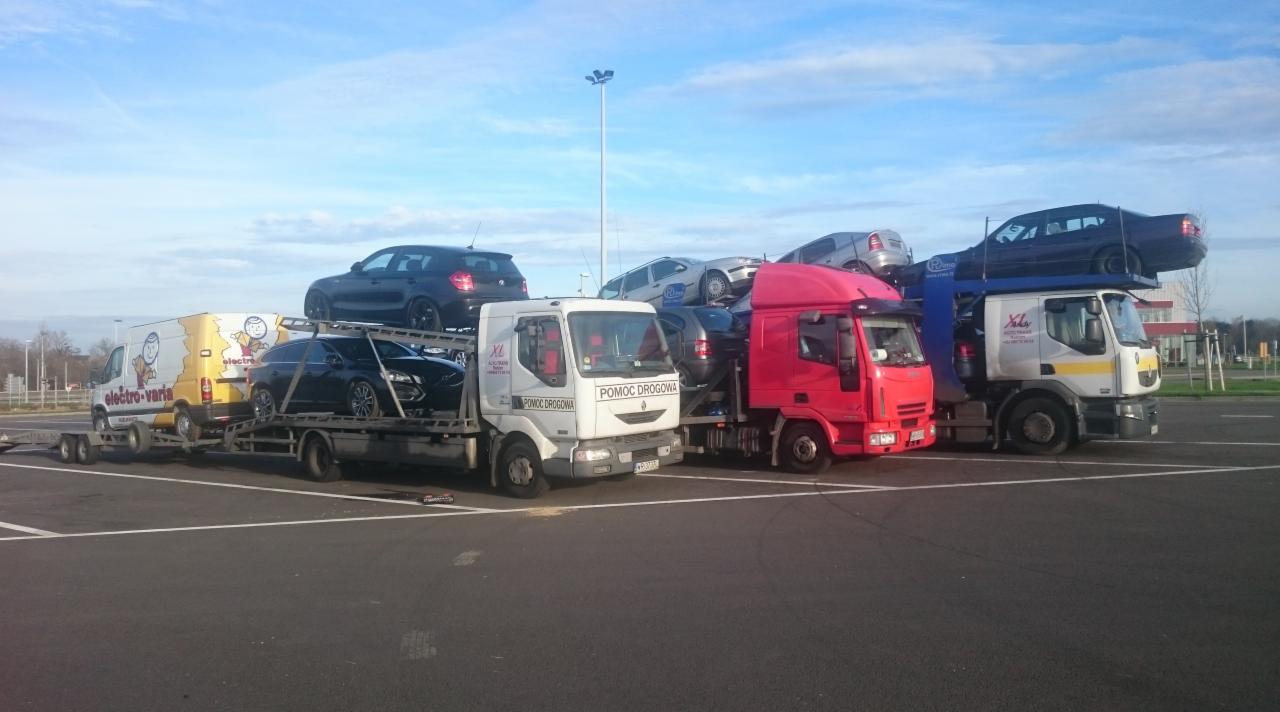 Trzy lawety i furgonetka transportują samochody osobowe na parkingu w słoneczny dzień.