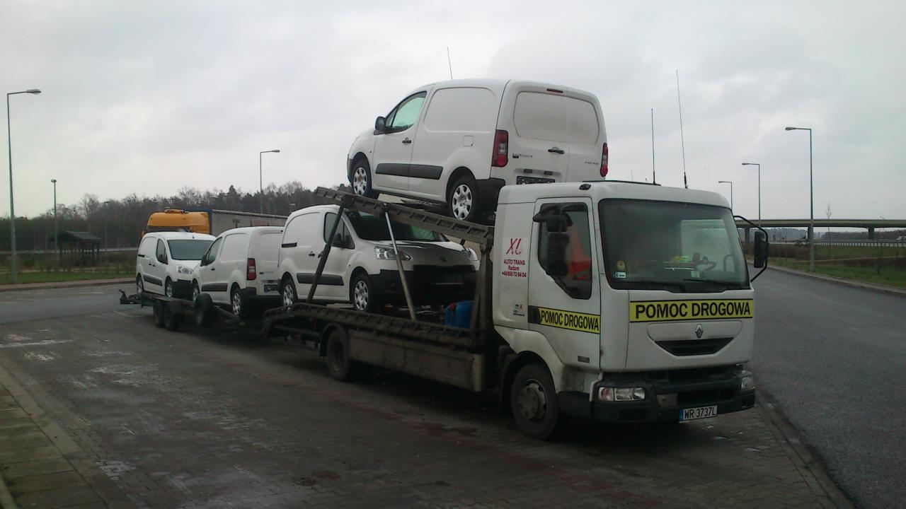 Laweta z napisem 'Pomoc Drogowa' przewozi trzy białe samochody dostawcze, jeden na przyczepie i jeden na dachu lawety, na tle pochmurnego nieba i krajobrazu.