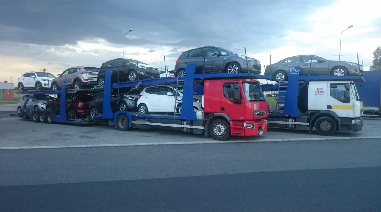 Dwa autotransportery wypełnione samochodami osobowymi różnych marek, stojące na parkingu pod pochmurnym niebem.