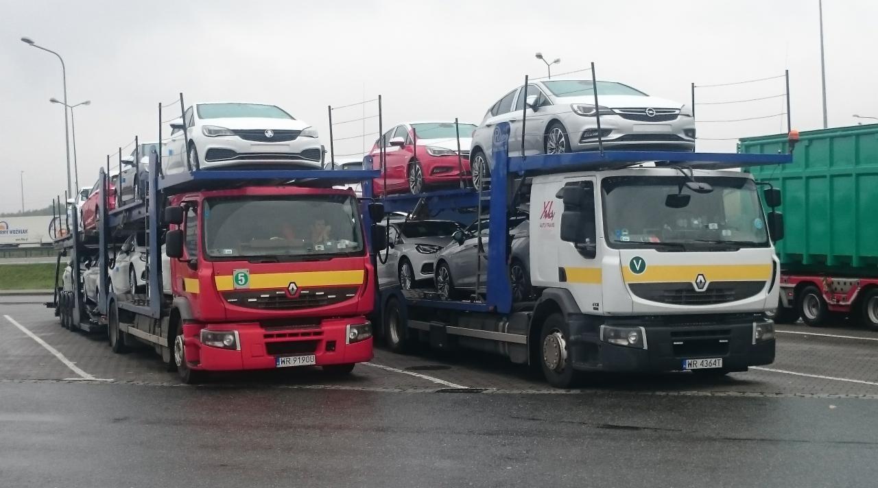 Dwa samochody ciężarowe typu laweta, jeden czerwony z numerem 5 na drzwiach, drugi biały z logo firmy XL AutoTrans, przewożące nowe samochody osobowe w różnych kolorach na parkingu.