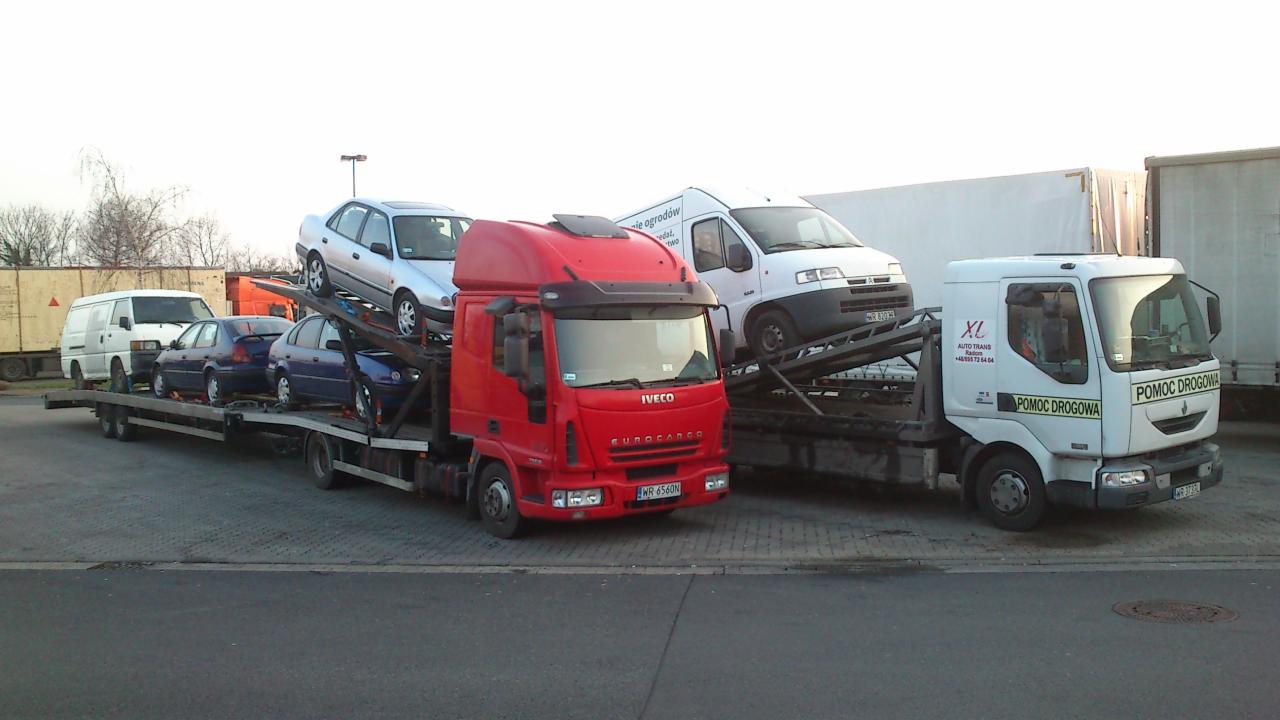 Dwa samochody transportowe, jeden czerwony Iveco Eurocargo i jeden biały Renault z napisem Pomoc Drogowa, załadowane różnymi pojazdami osobowymi i dostawczymi na parkingu.