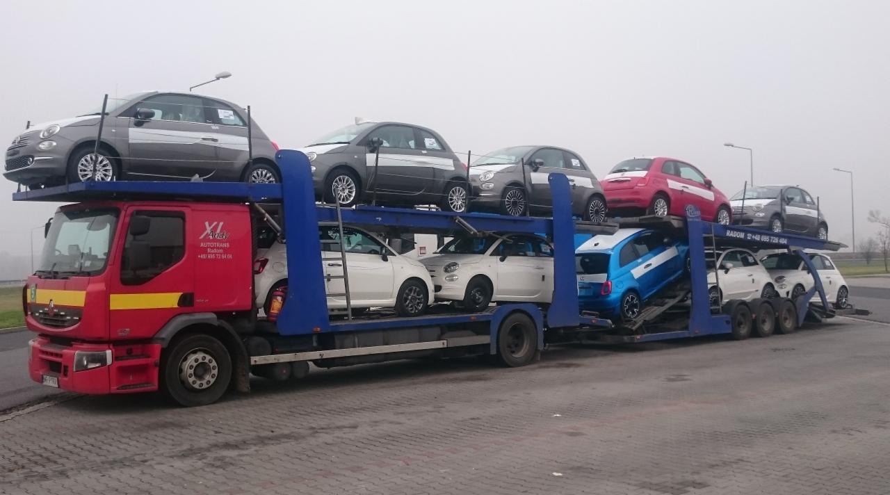 Czerwona laweta z niebieskimi elementami transportuje osiem samochodów Fiat 500 w różnych kolorach, zaparkowana na szarym parkingu w pochmurny dzień.