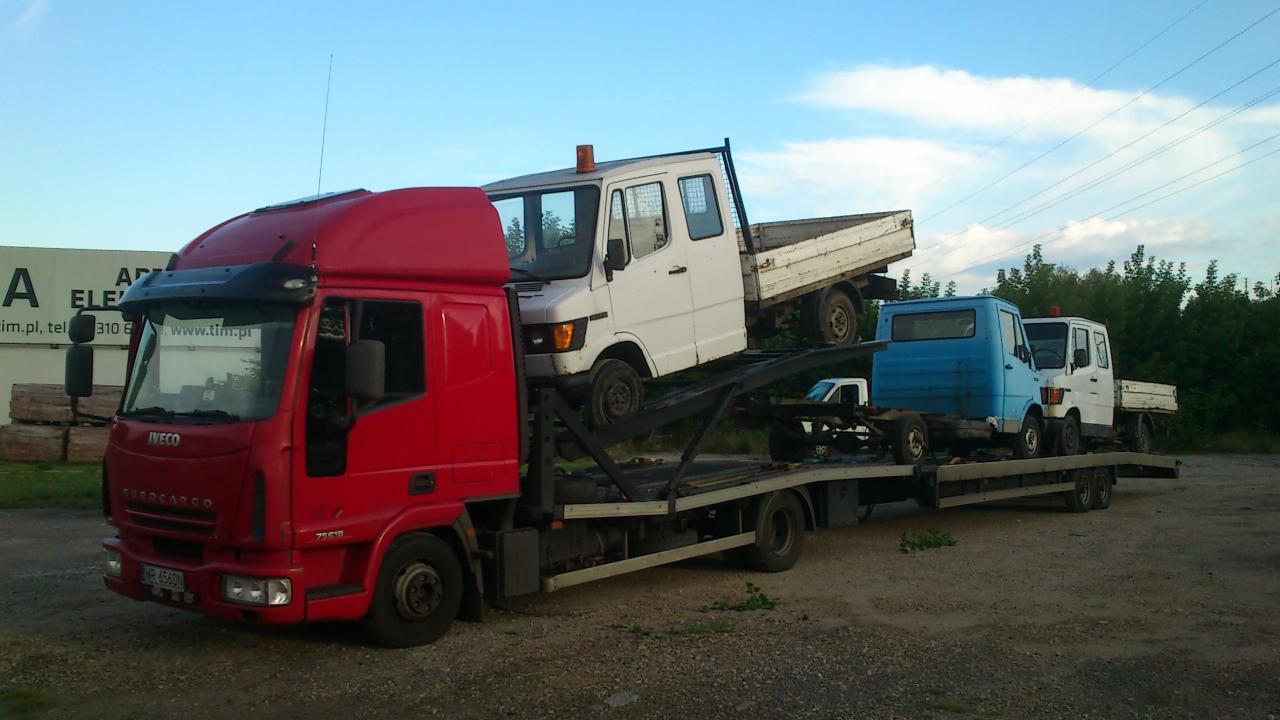 Czerwona laweta Iveco z trzema załadowanymi samochodami dostawczymi na platformie, zaparkowana na żwirowym placu w pochmurny dzień.