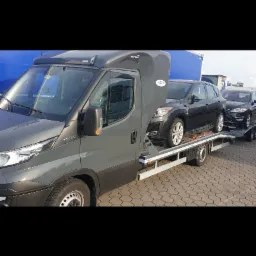 Szara laweta z dwoma samochodami osobowymi na platformie, widok z boku na tle niebieskiej naczepy.