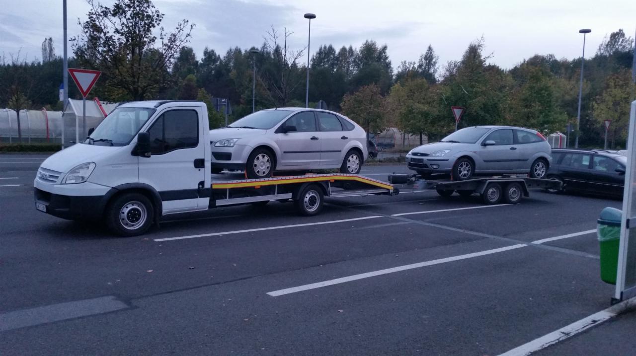 Biała laweta z dwoma srebrnymi Fordami Focus na parkingu, jeden na lawecie, drugi na przyczepie. Widoczne drzewa i znak drogowy.