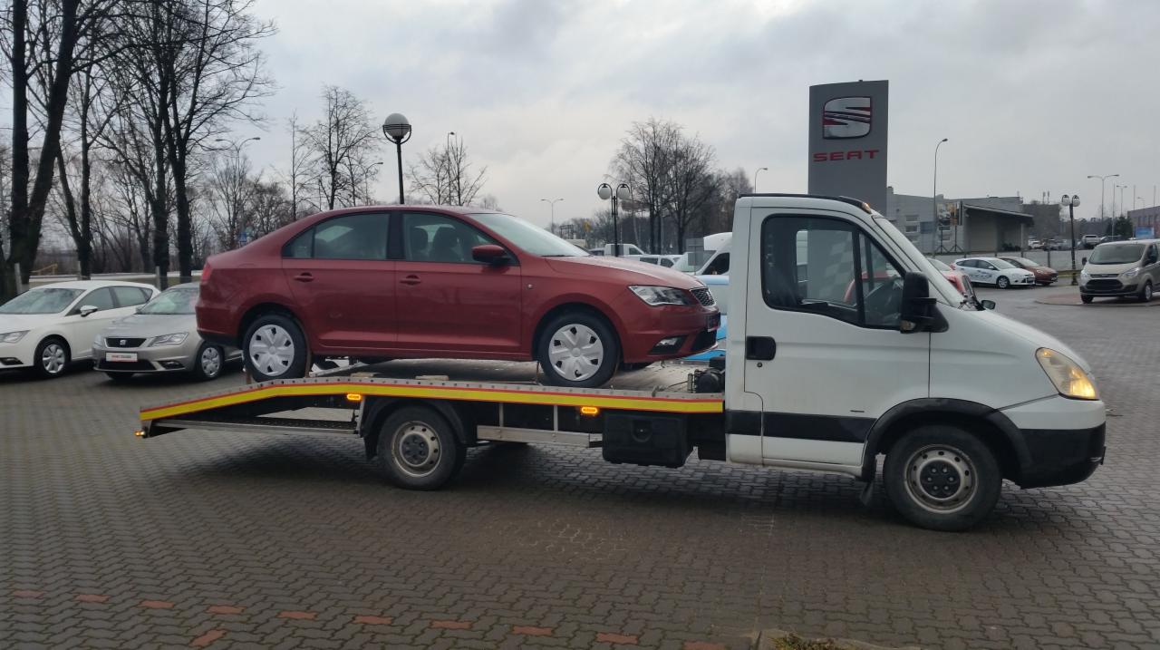 Czerwony Seat Toledo na lawecie przed salonem marki Seat. Biała laweta z żółtymi elementami. Szary dzień, kostka brukowa.