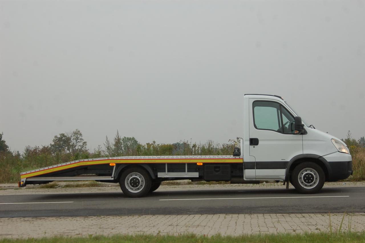 Biała laweta na drodze, widok z boku. Platforma z żółtymi akcentami, szare niebo w tle. Detal: brak ładunku na platformie.