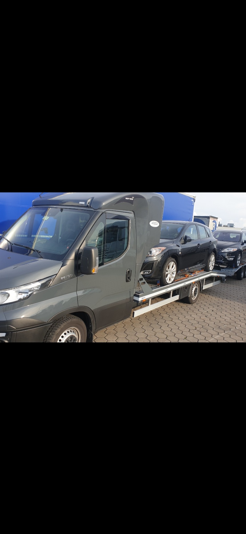 Szara laweta z dwoma samochodami osobowymi na platformie, widok z boku na tle niebieskiej naczepy.