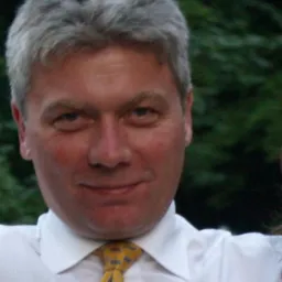 Portret mężczyzny z siwymi włosami, ubranego w białą koszulę i żółto-szary krawat, na tle zieleni. Uśmiechnięty, z widocznymi zmarszczkami wokół oczu.