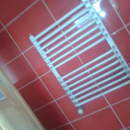 Biały grzejnik drabinkowy na ścianie z czerwonych płytek w łazience, widok z dołu. Widoczny fragment drewnianych drzwi.