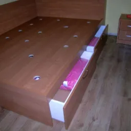 Drewniane łóżko z wysuwanymi szufladami na pościel, widoczna szafka nocna z lampką na blacie, podłoga z paneli.