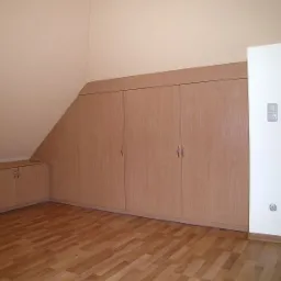 Zabudowa szafy wnękowej z jasnego drewna, dopasowana do skosu poddasza, z widocznymi drzwiczkami i drewnianą podłogą.