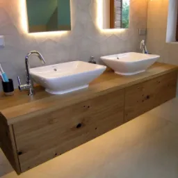 Łazienka w Kobylance z podwójną umywalką nablatową na szafce z blatem z jasnego drewna z widocznymi sękami i usłojeniami, oświetlona LED-ami od dołu i od góry, z szarą mozaikową ścianą w tle.