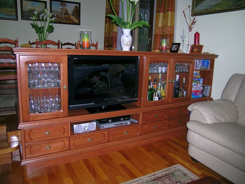 Drewniana komoda RTV z przeszklonymi witrynami, wypełniona szkłem i butelkami, z licznymi szufladami, na tle parkietu i jasnej sofy.