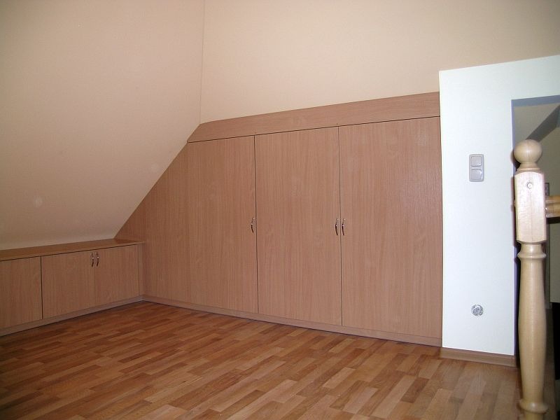 Zabudowa szafy wnękowej z jasnego drewna, dopasowana do skosu poddasza, z widocznymi drzwiczkami i drewnianą podłogą.